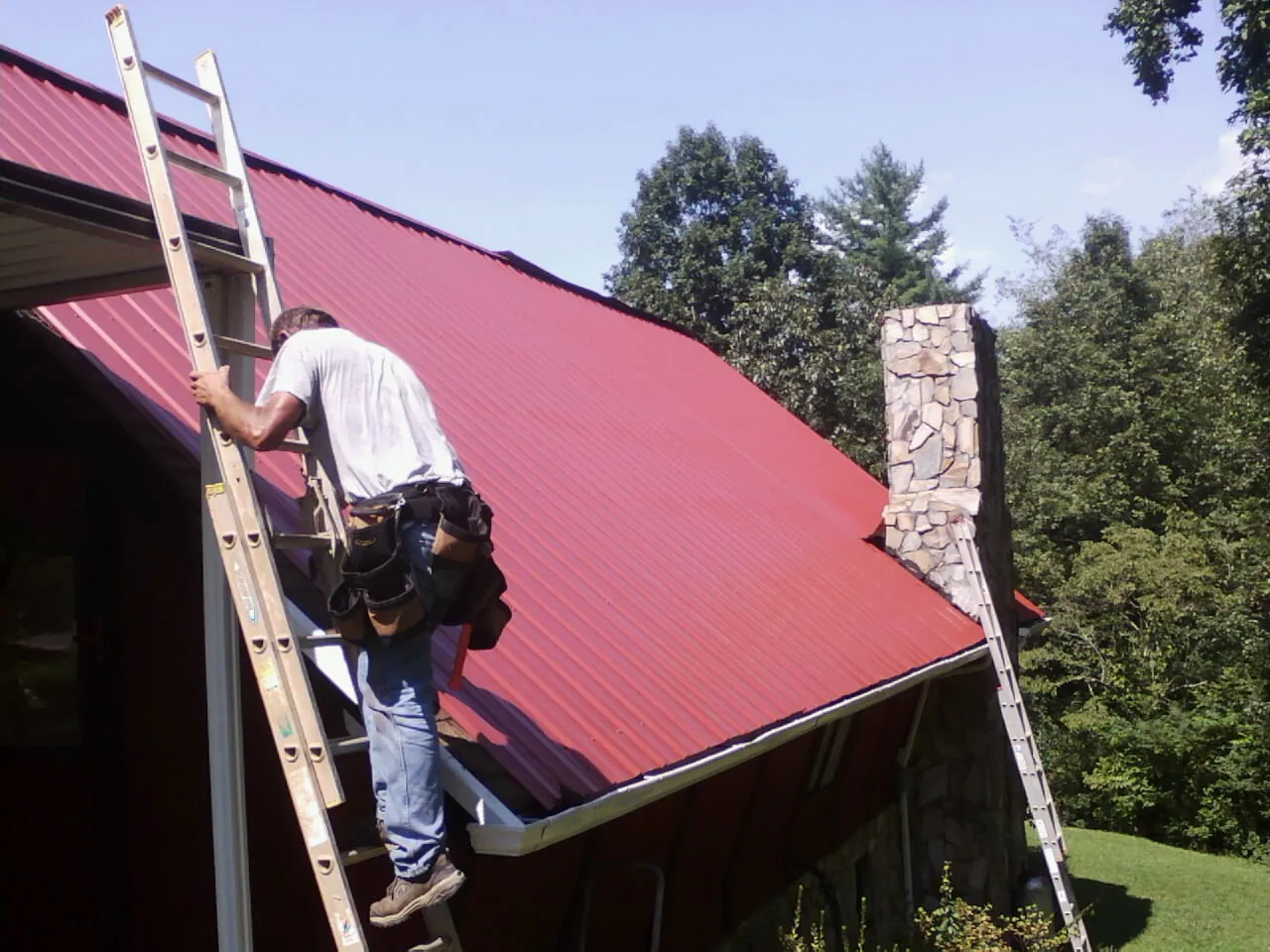 Roofer on a ladder beside a red metal roof and stone chimney — Soffit & Fascia Repair by Roofing Brothers in Douglass